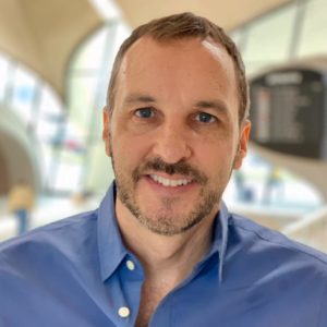 Jordan Friedman headshot photo, smiling man wearing light blue button up shirt in front of blurred background