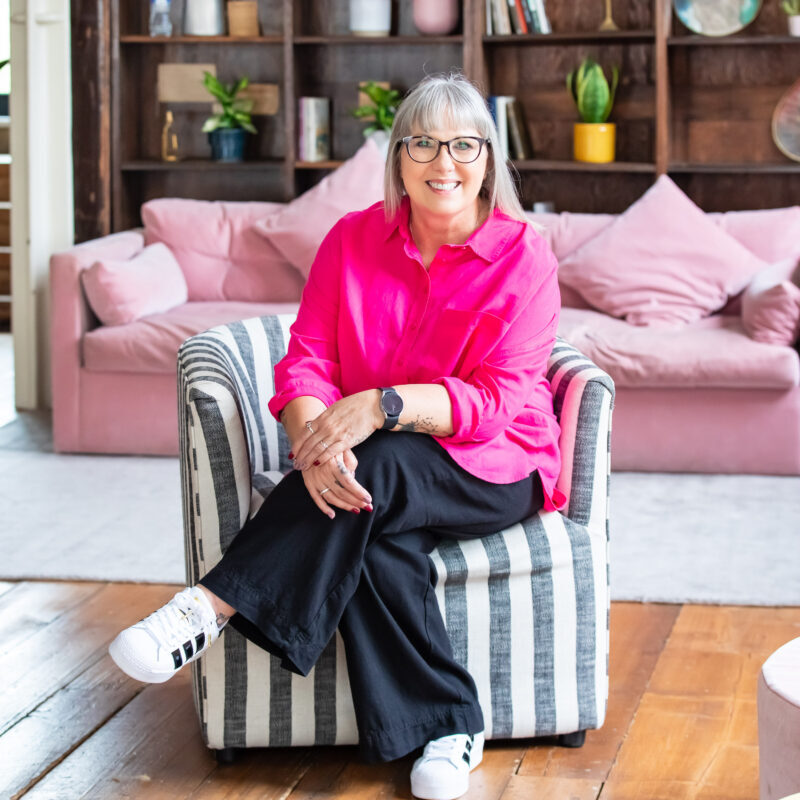 Debbie Green portrait headshot, smiling woman wearing a pink top black pants and white sneakers sitting on striped chair