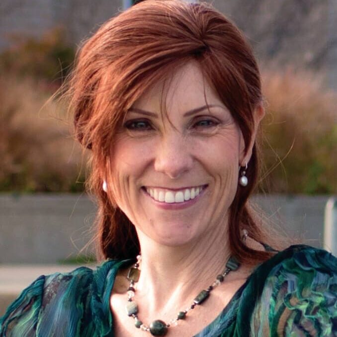 Ann Betz PCC headshot photo, smiling woman with red hair wearing cool toned top outside