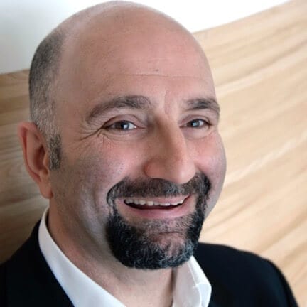 Aboodi Shabi PCC headshot photo, smiling man with dark beard wearing black blazer and white collared shirt in front of neutral background