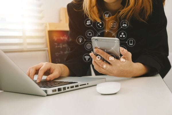 woman working on a laptop and holding a cellphone with an overlay of many icons around the cellphone