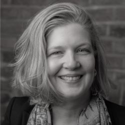 Liv Blaney ACC headshot photo, smiling woman with light hair wearing black blazer and scarf in front of brick wall black and white image