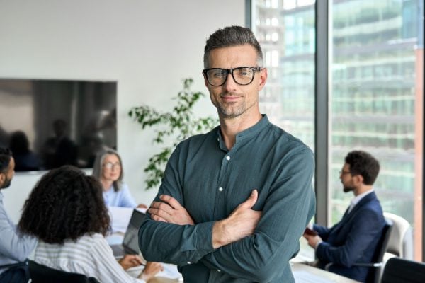 confident man smiling while others meet at a conference table behind him
