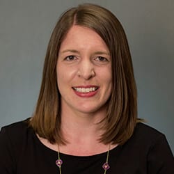 Abby Heverin headshot photo, smiling woman with brown hair and black top in front of gray background