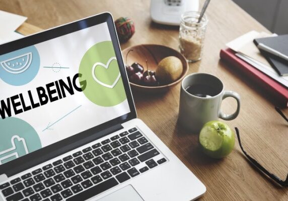 desk with laptop showing wellbeing graphic and apple coffee cup glasses and various other things on the desk