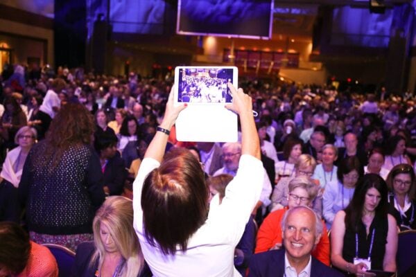 A woman in the foreground stands up and takes a photo of a diverse and inclusive crowd using her digital tablet, capturing the vibrancy of the community.