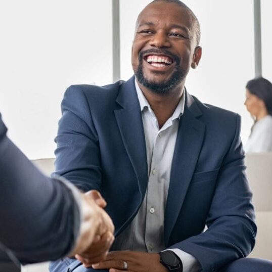 A smiling professional shaking hands with another professional in a light-filled, modern lounge.