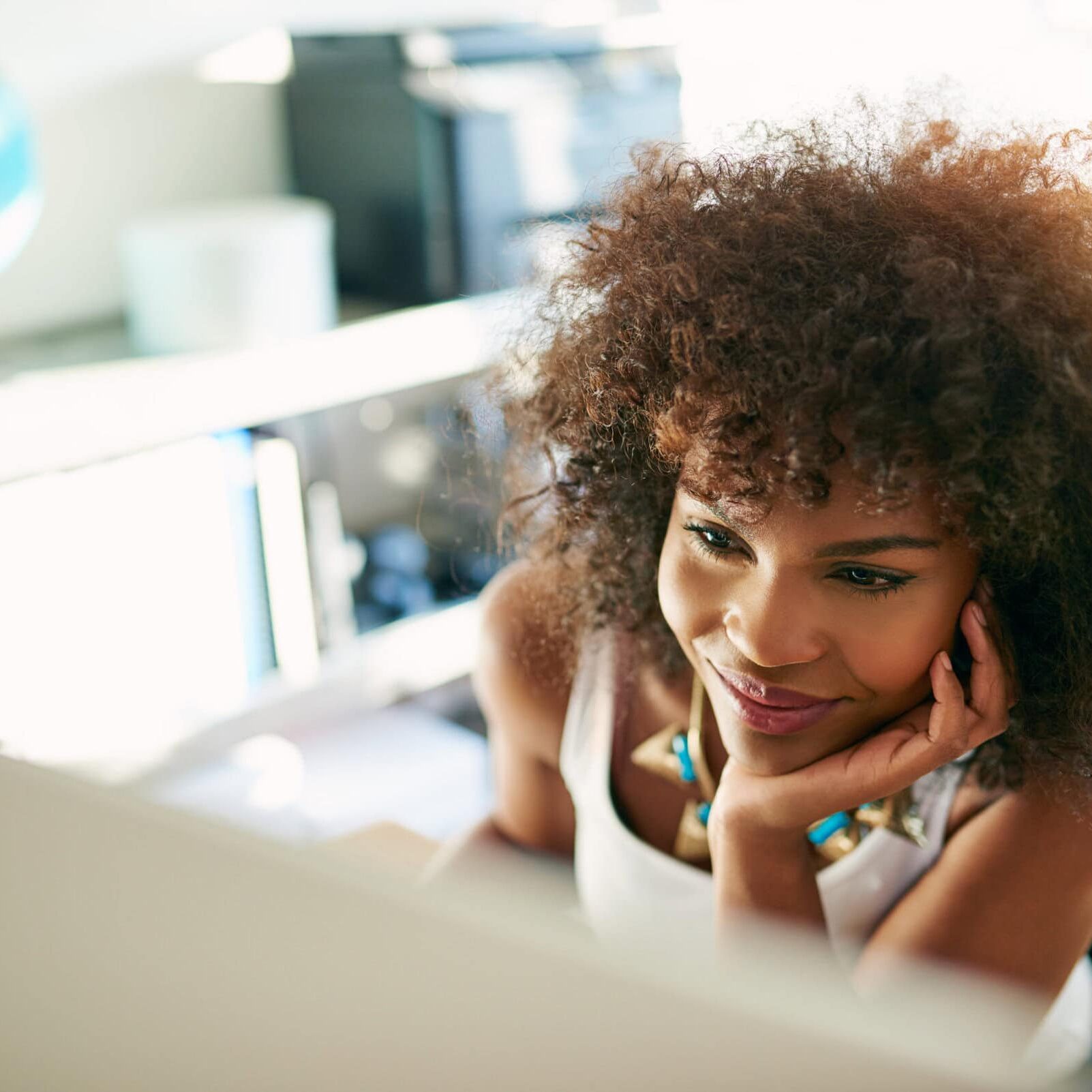 A young woman with curly hair smiling while working on coach professional development at a desk, with a globe and computer visible in a bright office setting.