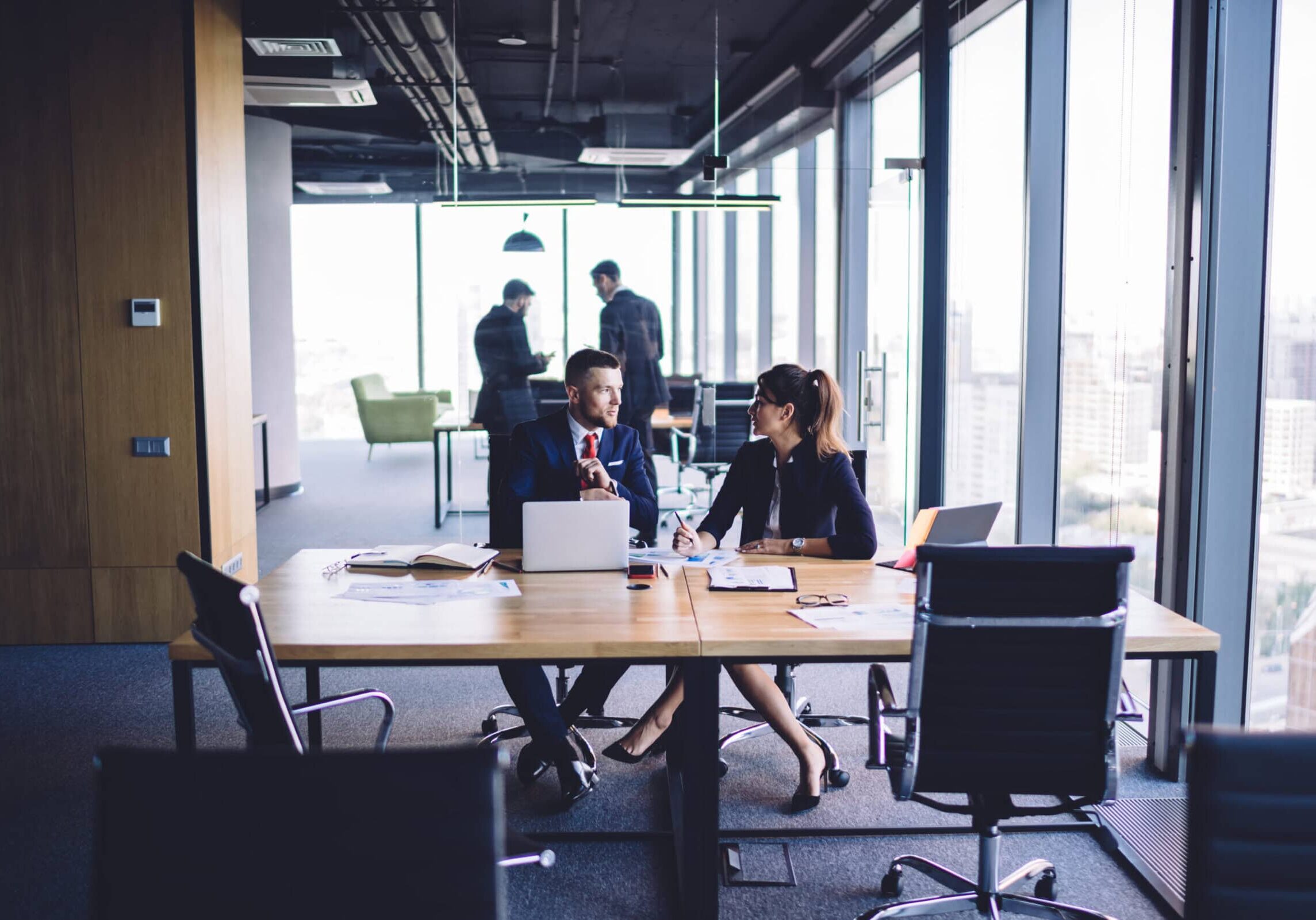 Two business professionals in a meeting at a table, discussing coaching. Two other professionals stand near windows overlooking a city.