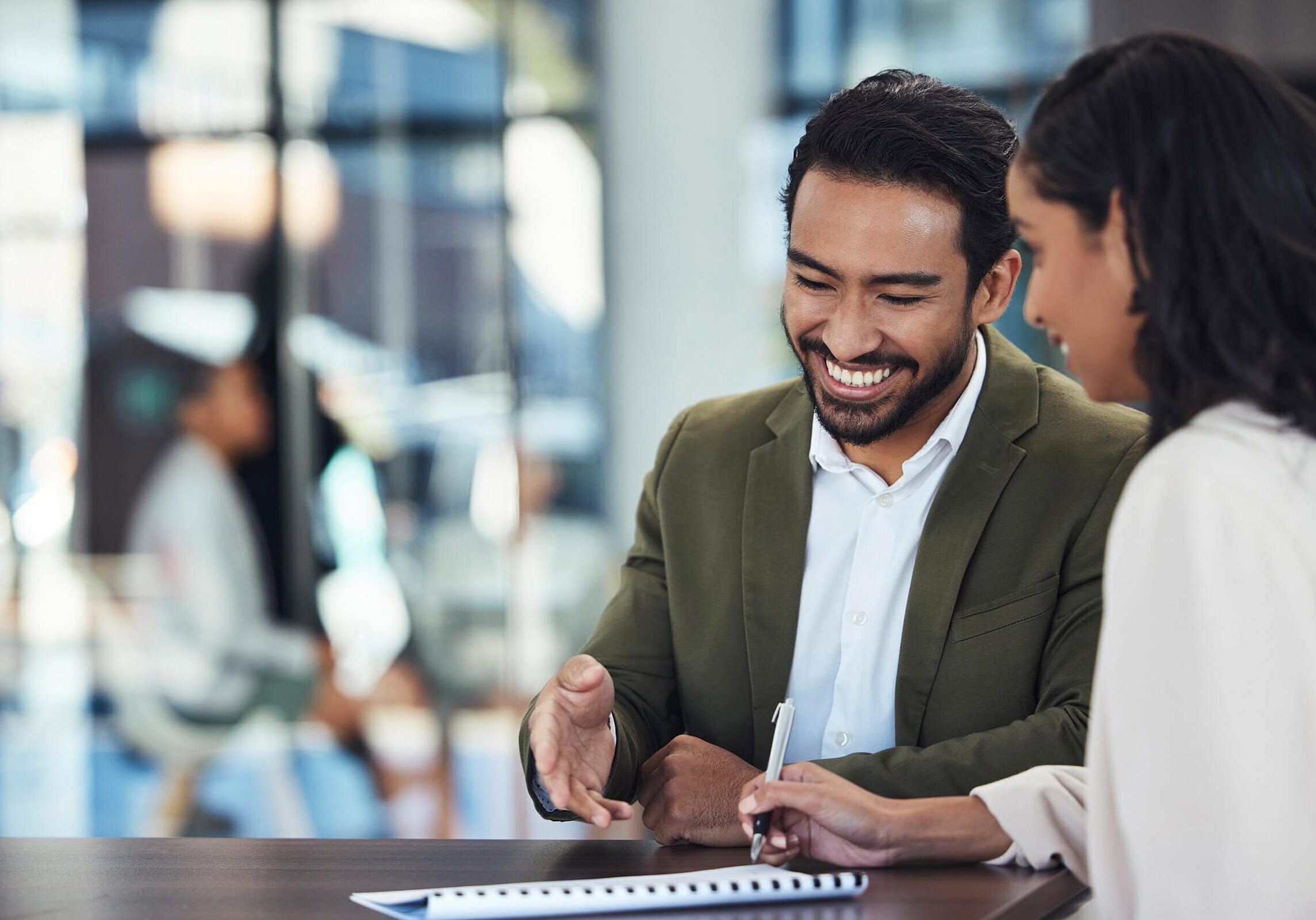 A man in a suit jacket smiling and gesturing while speaking with a woman in a business setting, symbolizing coaching partnerships.
