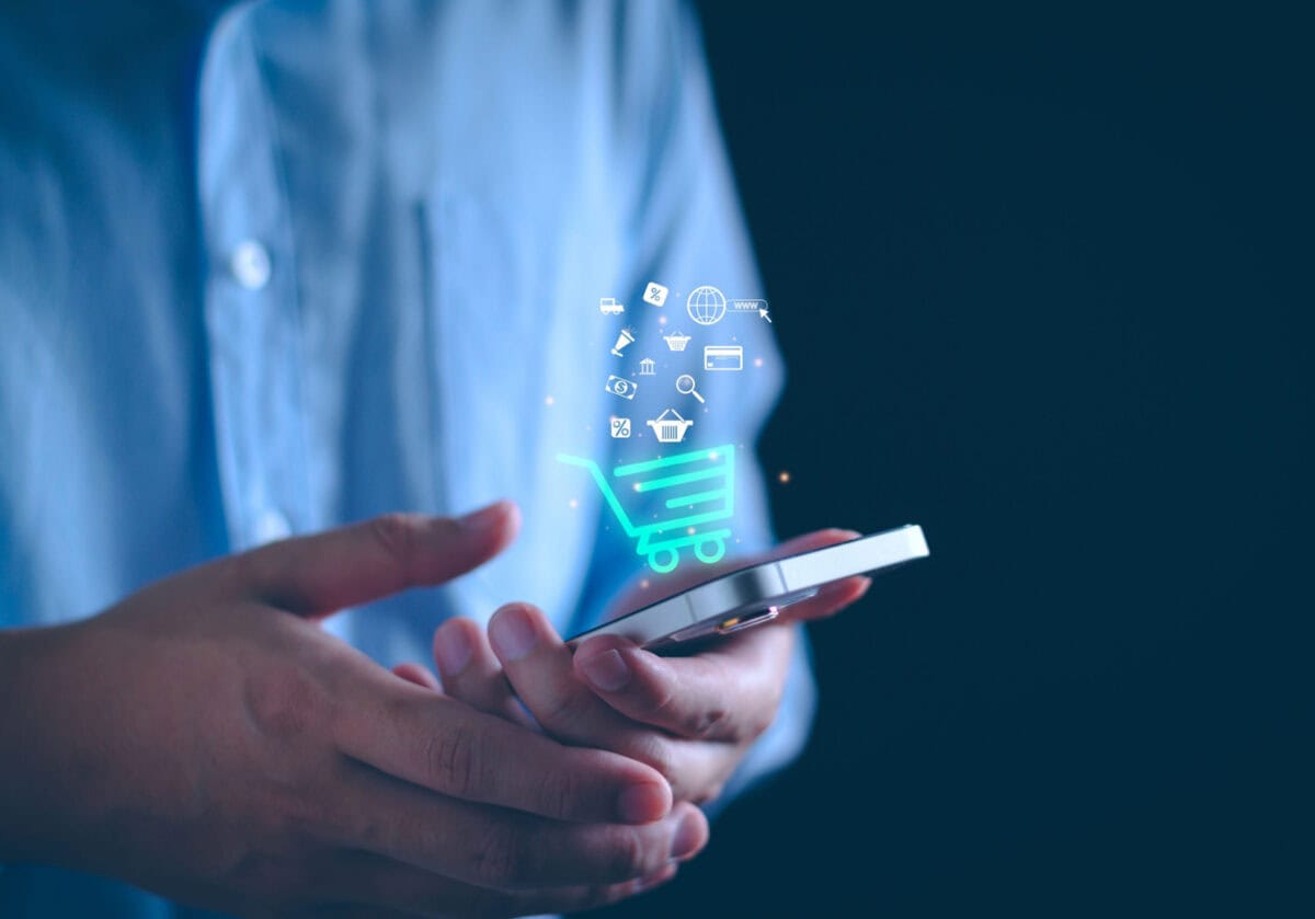 A man in a blue dress shirt holds a smartphone with a floating shopping cart icon above it.