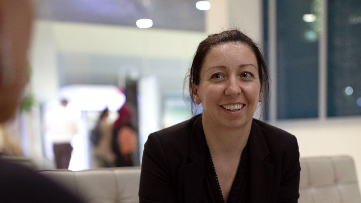 Jenn Header, woman with brown hair smiling wearing dark top sitting on a couch