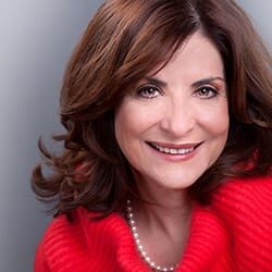 Marcia Reynolds MCC headshot photo, smiling woman with brown hair wearing a string of pearls and red sweater in front of gray wall