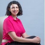 Jihane Labib headshot photo, smiling woman wearing pink top with hands clasped on her knee sitting in front of light background