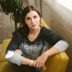 Michelle Rockwood ACC Headshot photo, dark-haired woman sitting on a yellow couch next to a wall with a plant in the background