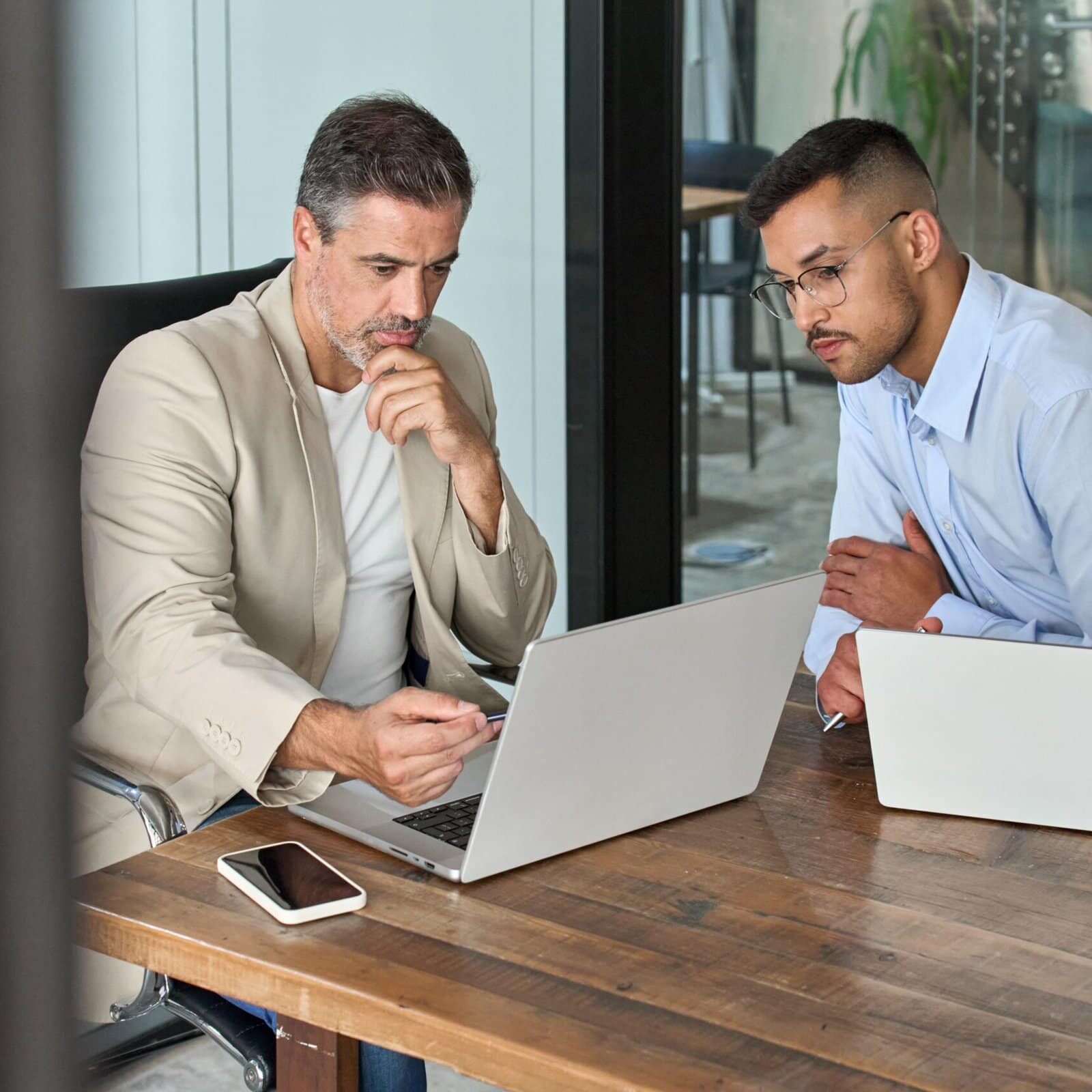 two men in front of their respective laptops one showing the other something on his laptop