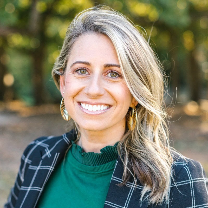 Traci Stormie ACC headshot photo, smiling woman with blond hair wearing gold earrings, a green shirt and black blazer outside
